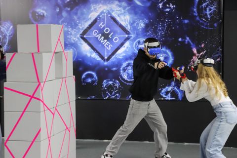 Zwei Personen spielen ein Virtual-Reality-Spiel in einer modernen Umgebung. Im Hintergrund sind leuchtende Grafiken und eine Struktur aus Kartons mit pinken Markierungen zu sehen.