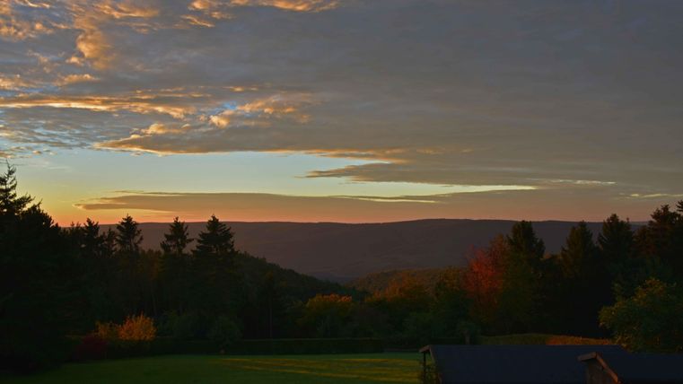 Eine malerische Landschaft bei Sonnenuntergang mit sanften Hügeln und buntem Himmel. Im Vordergrund sind grüne Wiesen und einige Bäume zu sehen.
