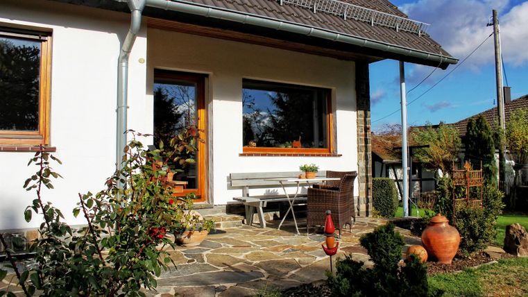 Ein modernes Haus mit einem einladenden Eingangsbereich. Vor der Haustür stehen ein Gartenstuhl und Töpfe, umgeben von Pflanzen.