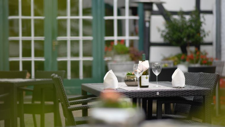 Ein gemütlicher Außenbereich eines Restaurants mit einem gedeckten Tisch. Im Hintergrund sind grüne Fenster und blühende Pflanzen zu sehen.