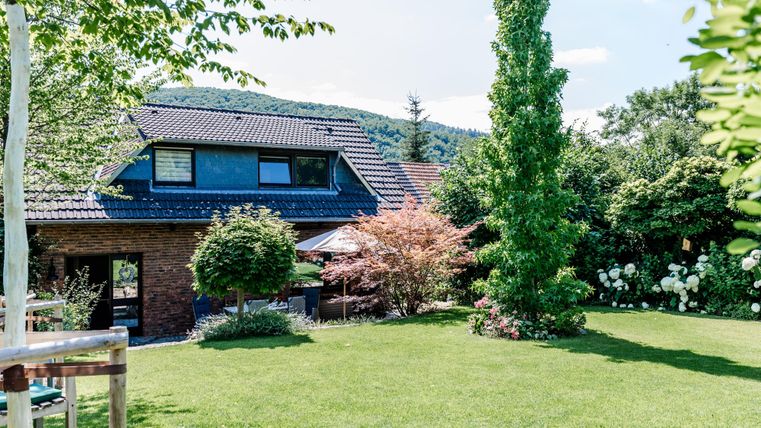 Ein schönes Haus umgeben von einem gepflegten Garten. Die Landschaft ist grün mit Bäumen und blühenden Pflanzen.