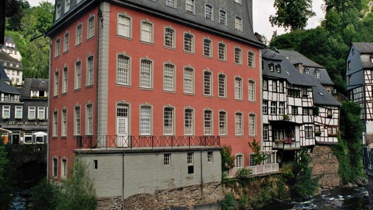 Ein historisches Gebäude in roter Farbe, umgeben von alten Fachwerkhäusern. Im Vordergrund fließt ein kleiner Fluss.