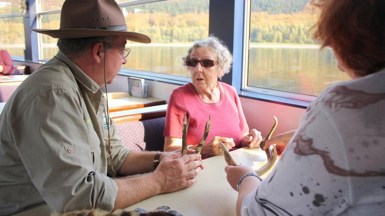 Eine Gruppe von drei Personen sitzt an einem Tisch in einem Boot. Sie diskutieren und halten Felle und Gegenstände in den Händen, während die Landschaft im Hintergrund sichtbar ist.