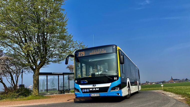 Ein Stadtbus fährt an einer Haltestelle vorbei, umgeben von einer grünen Landschaft. Der Himmel ist klar und blau.