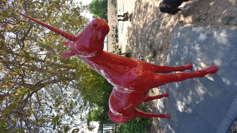 Ein rotes Einhorn aus Kunststoff steht auf einem Gehweg. Im Hintergrund sind Menschen und Hunde auf einem Spielplatz zu sehen.