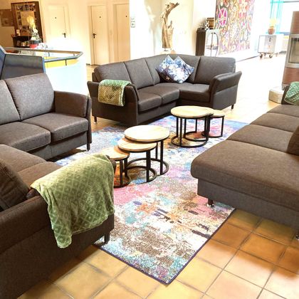 Eine gemütliche Sitzecke mit mehreren grauen Sofas und Couchtischen. Der Boden ist mit einem bunten Teppich dekoriert, und eine grüne Decke liegt auf einem Sofa.