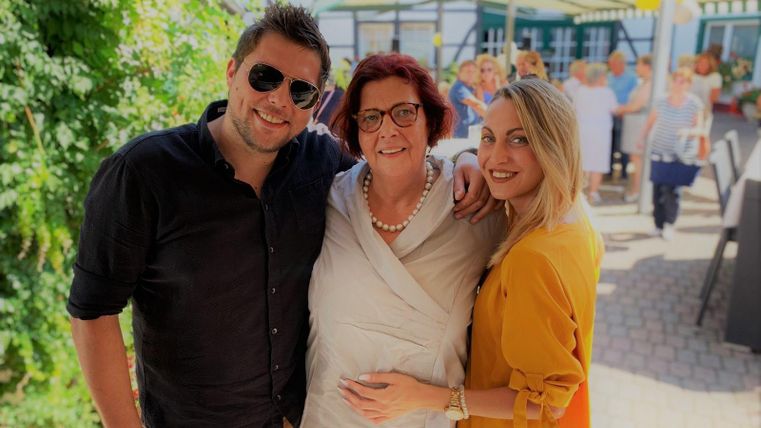 Drei Personen stehen zusammen und lächeln in die Kamera. Im Hintergrund sind weitere Menschen und eine sonnige Terrasse zu sehen.
