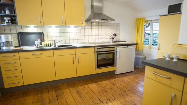 Eine moderne Küche mit gelben Schränken und Holzfußboden. Sie ist gut ausgestattet und hat große Fenster mit Vorhängen.