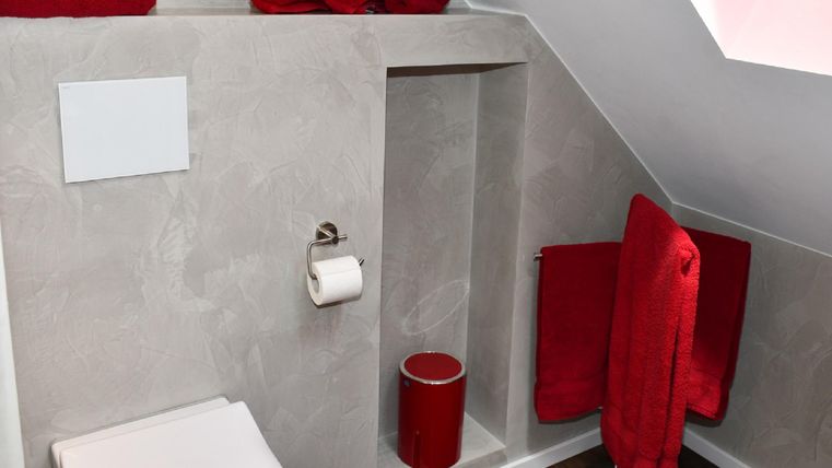 Ein modernes Badezimmer mit einer schrägen Decke und grauen Wänden. Rote Handtücher und ein roter Becher setzen farbliche Akzente.