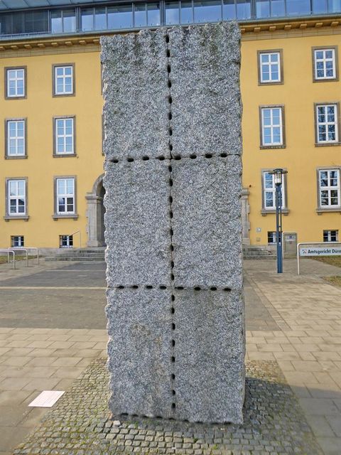 Ein großer, rechteckiger Steinsockel mit strukturierten Oberflächen steht vor einem gelben Gebäude. Das Design enthält eine Reihe von Löchern, die ein interessantes Muster bilden.