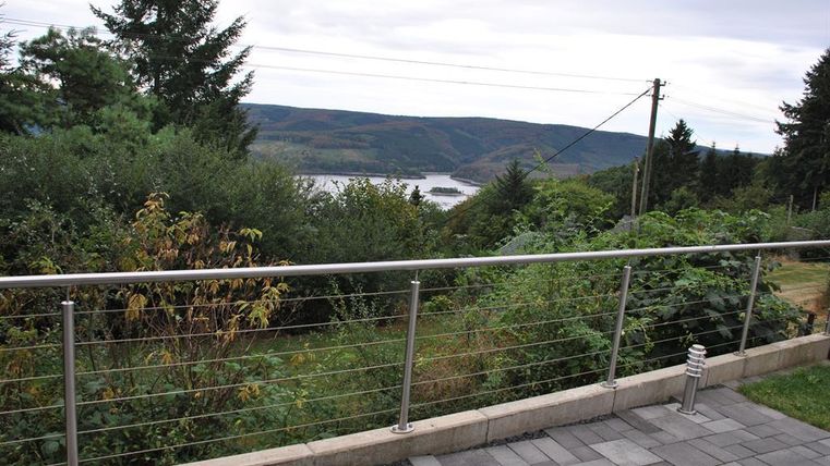 Eine schöne Aussicht über grüne Hügel und einen Fluss. Im Vordergrund befindet sich ein moderner Geländer und eine Pflasterfläche.