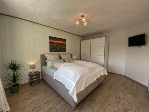 Ein gemütliches Schlafzimmer mit einem großen Bett und hellen Kissen. An der Wand hängt ein Bild und im Raum steht ein Fernseher sowie eine Pflanze.