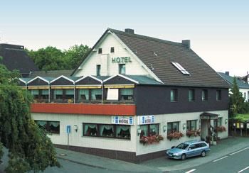 Ein Hotelgebäude mit einem geschützten Außenbereich und einem klaren Himmel. Vor dem Hotel steht ein blauer Wagen und es gibt Bäume in der Umgebung.