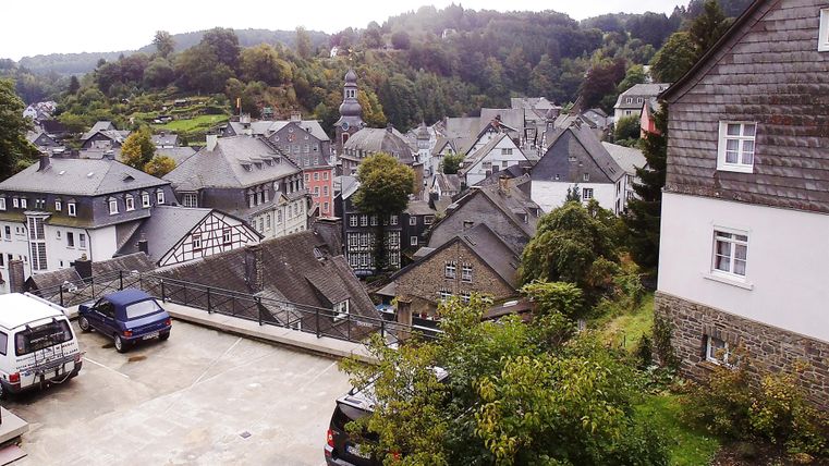 Eine malerische Stadtansicht mit traditionellen Häusern und dichten Bäumen. Im Hintergrund sind Hügel und eine Kirche zu sehen.