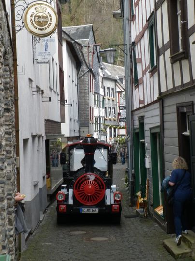 Eine enge, charmante Gasse mit historischen Gebäuden und einem bunten Zug, der durch die Straße fährt. Im Hintergrund sieht man eine Person, die die Umgebung betrachtet.