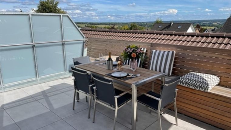 Eine gemütliche Terrasse mit einem Esstisch und Stühlen. Auf dem Tisch stehen Blumen und eine Flasche Wein, mit Blick auf die Landschaft.