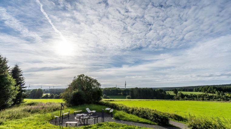 Eine weite, grüne Wiese unter einem klaren Himmel mit vielen Wolken. Im Vordergrund stehen Sitzgelegenheiten, ideal zum Entspannen und die Aussicht zu genießen.