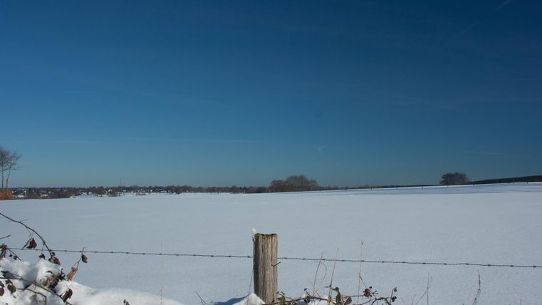 Eine schneebedeckte Landschaft unter einem klaren blauen Himmel. Im Vordergrund ist ein Holzpfosten mit einem Zaun zu sehen.