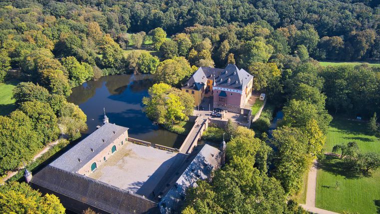 Ein malerisches Herrenhaus umgeben von Bäumen und einem ruhigen Teich. Die Aufnahme zeigt die weitläufige Landschaft und die Architektur des Gebäudes.