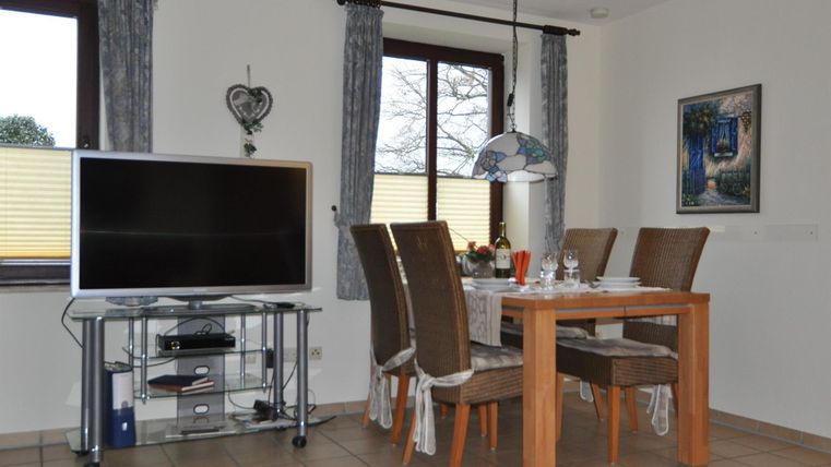 Ein moderner Essbereich mit einem Holztisch und gepolsterten Stühlen. Ein großer Fernseher steht auf einem Tisch, und durch die Fenster fällt natürliches Licht.