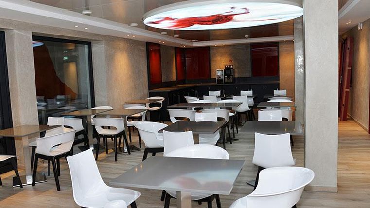 Ein modernes Restaurant mit weißen Tischen und Stühlen. Die Wände sind hell und es gibt große Fenster, die Tageslicht hereinlassen.