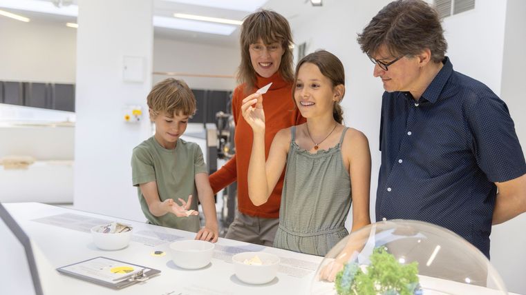 Eine Familie steht an einem Tisch in einem modernen Ausstellungsraum. Sie schauen interessiert auf Objekte und tauschen sich aus.