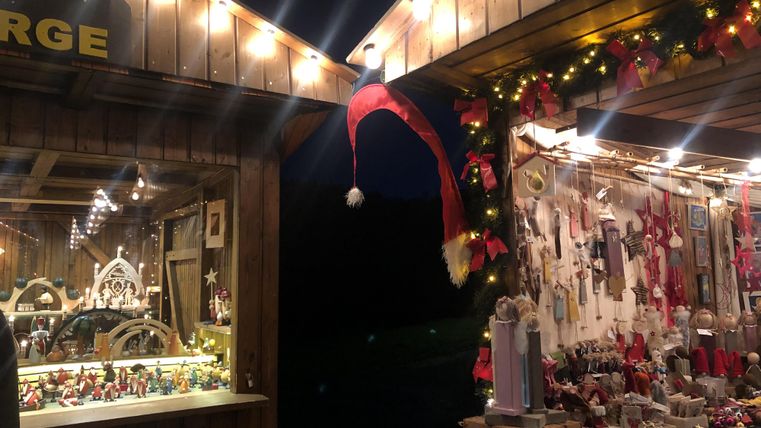 Ein festlich dekorierter Weihnachtsmarktstand mit bunten Lichtern und handgemachten Geschenken. Im Hintergrund hängt ein rotes Weihnachtsmützen-Dekor.
