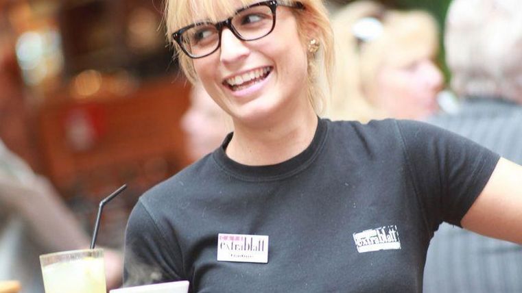 Eine freundliche Bedienung mit Brille lächelt in einem Restaurant. Im Hintergrund sind weitere Gäste zu sehen.