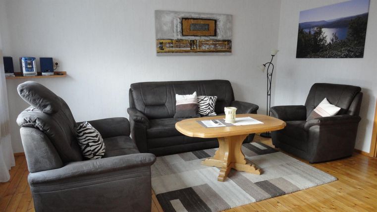 Ein gemütliches Wohnzimmer mit einer grauen Couchgarnitur und einem runden Holztisch. An der Wand hängt ein Bild und ein kleines Regal ist mit Lautsprechern dekoriert.