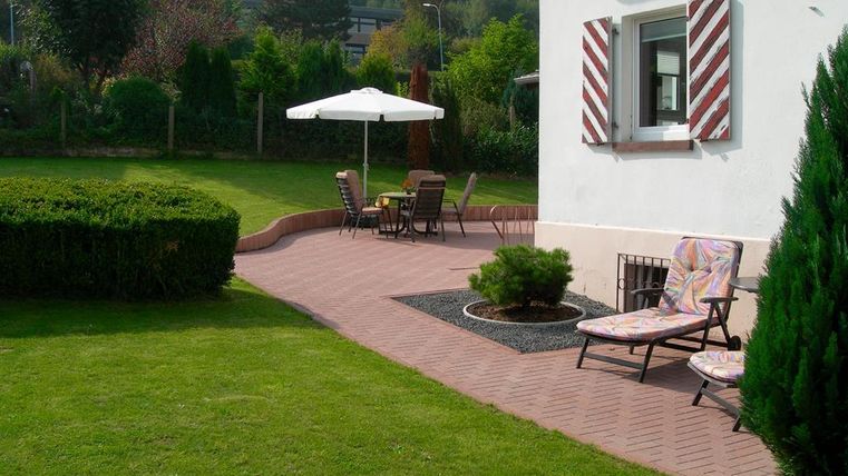 Ein schöner Garten mit einer Terrasse und Gartenmöbeln. Im Hintergrund steht ein Sonnenschirm und eine gepflegte Rasenfläche.