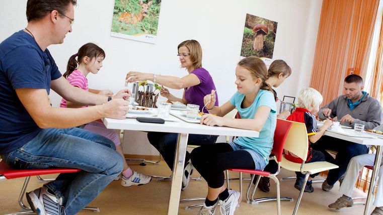Eine Gruppe von Kindern und Erwachsenen sitzt an einem Tisch und malt. Es herrscht eine kreative Atmosphäre in einem hellen Raum.