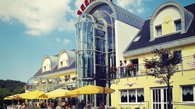 Ein modernes Restaurant mit einer großen Glasfassade und gelben Sonnenschirmen. Im Vordergrund sind Gäste, die draußen sitzen und essen.