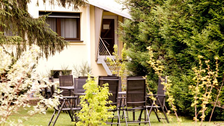 Ein gepflegter Garten mit einem Set aus Stühlen und Tischen. Im Hintergrund ist ein Haus und hohe Pflanzen zu sehen.