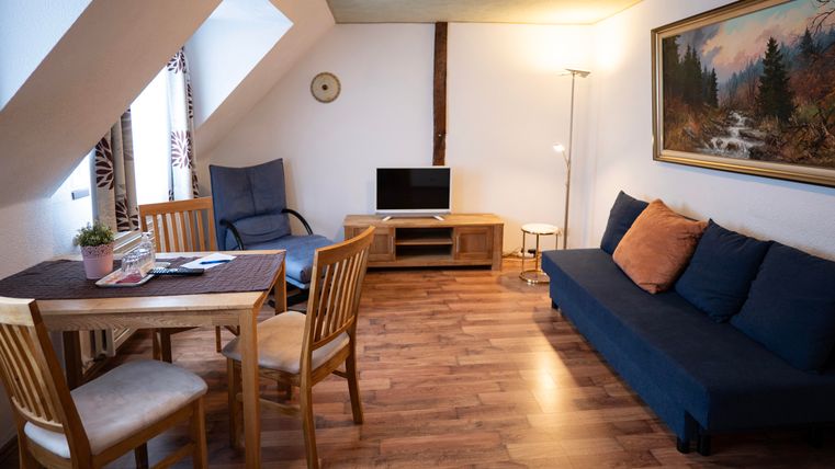 Ein gemütliches Wohnzimmer mit einem blauen Sofa, einem Esstisch und einem Fernseher. Die Wände sind hell, und es gibt ein Fenster mit Vorhängen.