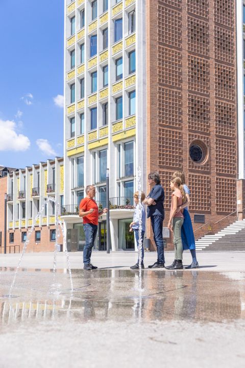 Eine Gruppe von vier Personen steht vor einem modernen Gebäude an einem sonnigen Tag. Der Platz ist nass, vermutlich von einem Springbrunnen.