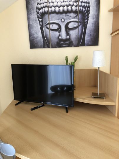 Ein moderner Fernseher steht auf einem hellen Holztisch. Im Hintergrund hängt ein Bild eines Buddha-Gesichts.