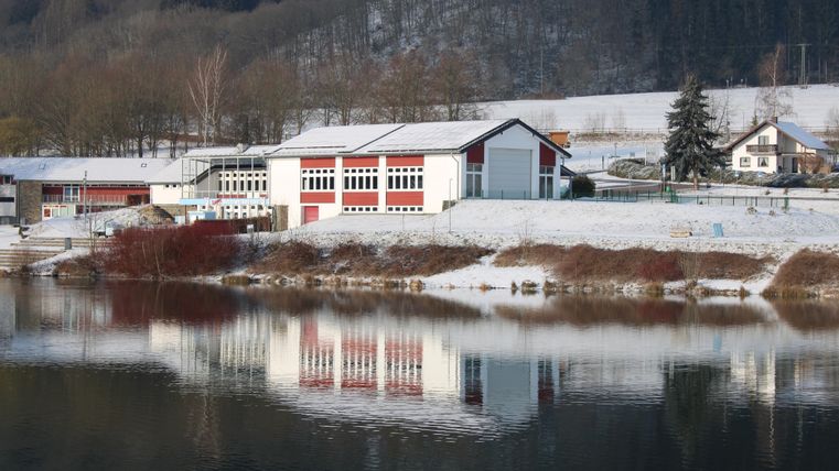 Ein modernes Gebäude am Ufer eines ruhigen Sees, umgeben von Schnee. Die Landschaft ist klar und spiegelt sich im Wasser wider.