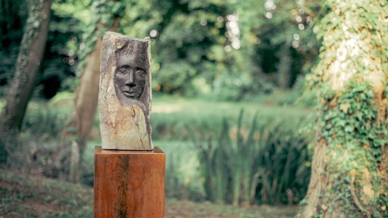 Eine Holzskulptur mit einem Gesicht steht auf einem Podest in einem grünen Park. Im Hintergrund sind Bäume und eine ruhige Landschaft zu sehen.