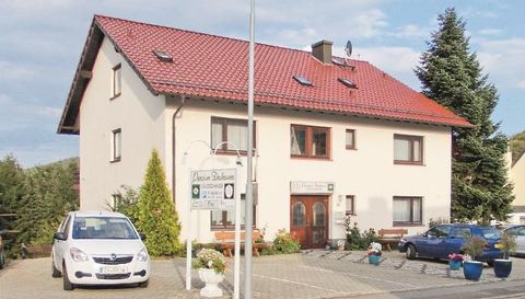 Ein zweistöckiges Wohnhaus mit rotem Dach und mehreren Fenstern. Vor dem Haus stehen Fahrzeuge und blühende Pflanzen in Töpfen.