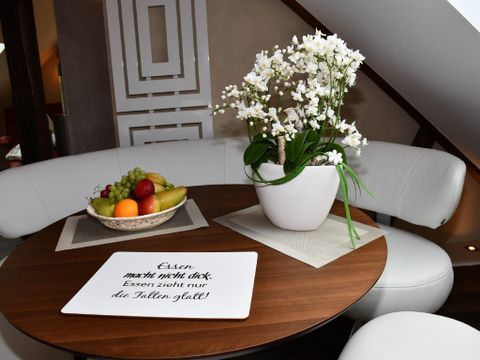 Ein stilvoller Tisch mit einer Schale voller frischer Früchte und einer blühenden Pflanze. Auf der Tischplatte liegt ein Schild mit der Aufschrift: "Essen macht nicht dick, Essen zählt nur das Talent gut!"