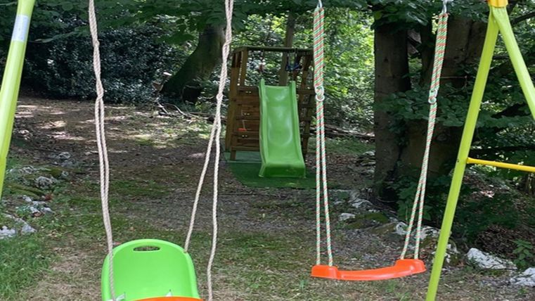 Eine Kinderschaukel mit zwei Sitzen steht im Grünen. Im Hintergrund ist eine Rutsche zu sehen.