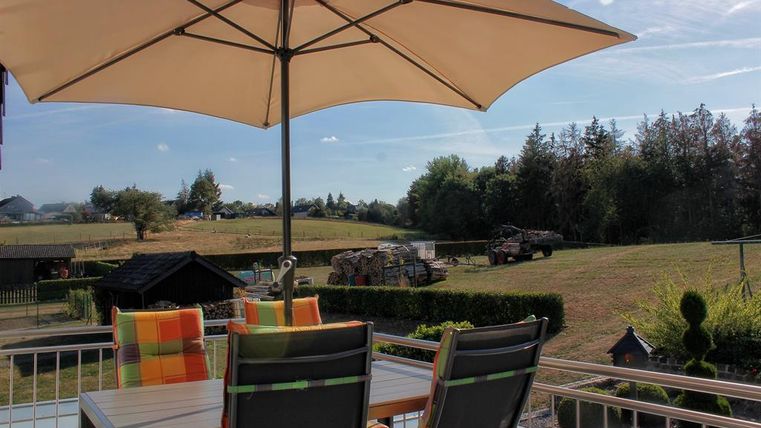 Ein gemütlicher Balkon mit einem Sonnenschirm und einem Tisch. Im Hintergrund sieht man eine grüne Wiese und Bäume.