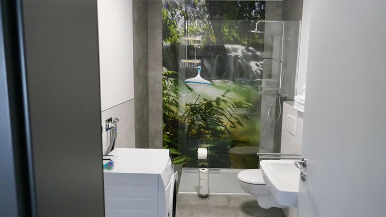Ein modernes Badezimmer mit einer Waschmaschine und einer hellen, einladenden Umgebung. Eine Fototapete mit einem schönen Wasserfall sorgt für eine entspannende Atmosphäre.