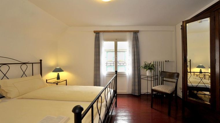 Ein helles Schlafzimmer mit einem Doppelbett und einem großen Fenster. Der Raum ist schlicht und gemütlich eingerichtet, mit einem Tisch und einem Spiegel.