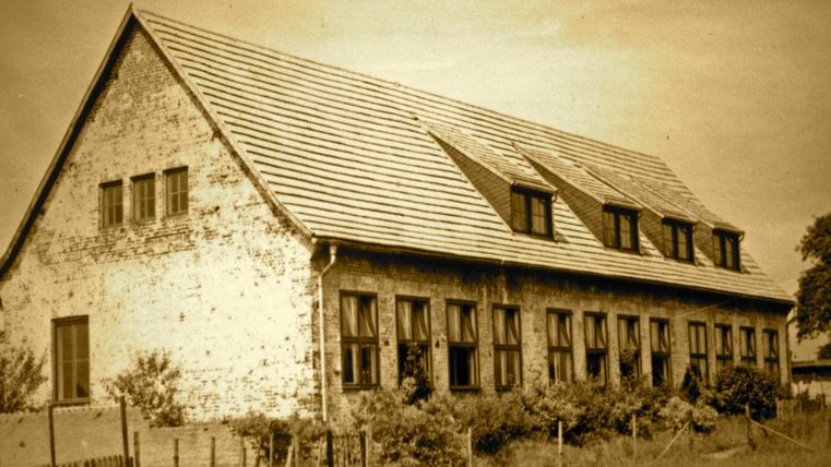 Ein altes, rustikales Gebäude mit einem geneigten Dach und mehreren Fenstern. Das Bild hat einen sepiafarbenen Look, der einem nostalgischen Gefühl verleiht.
