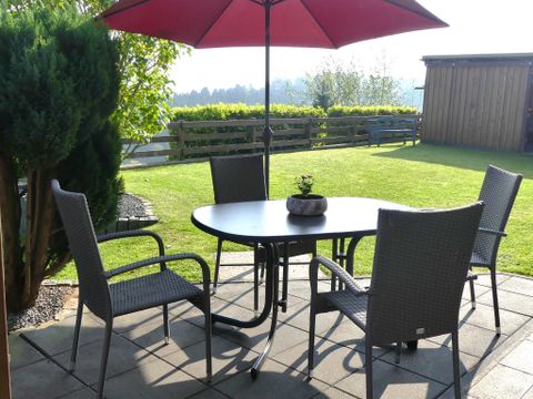 Ein einladender Außenbereich mit einem runden Tisch und vier Stühlen unter einem roten Sonnenschirm. Der Garten ist grün und gepflegt, ideal für entspannte Stunden im Freien.