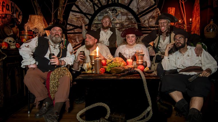 Eine Gruppe von Piraten sitzt um einen Tisch mit Lebensmitteln und Getränken. Die Atmosphäre ist festlich und die Umgebung ist im Piratenstil dekoriert.