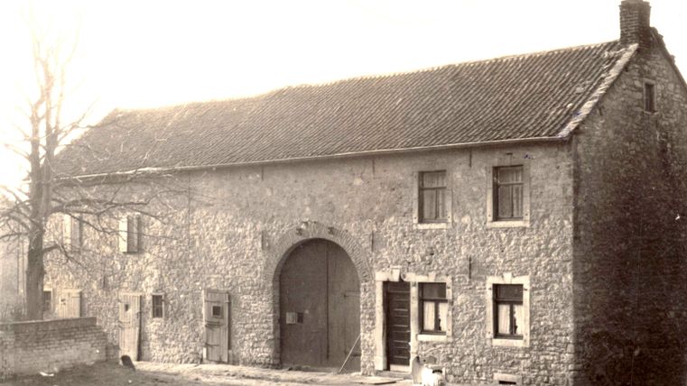 Ein altes Steinhaus mit einem großen Tor und mehreren Fenstern. Im Vordergrund sind einige Bäume und eine klare Straße zu sehen.