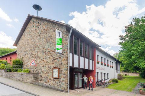 Ein modernes Gebäude mit einer Steinfassade und großen Fenstern. Vor dem Eingang stehen Fahrräder und es gibt grüne Pflanzen in der Nähe.