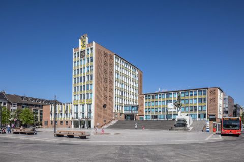 Ein modernes Gebäude mit einer markanten Fassade steht auf einem Platz. Die klare blaue Himmel gibt dem Ort eine einladende Atmosphäre.